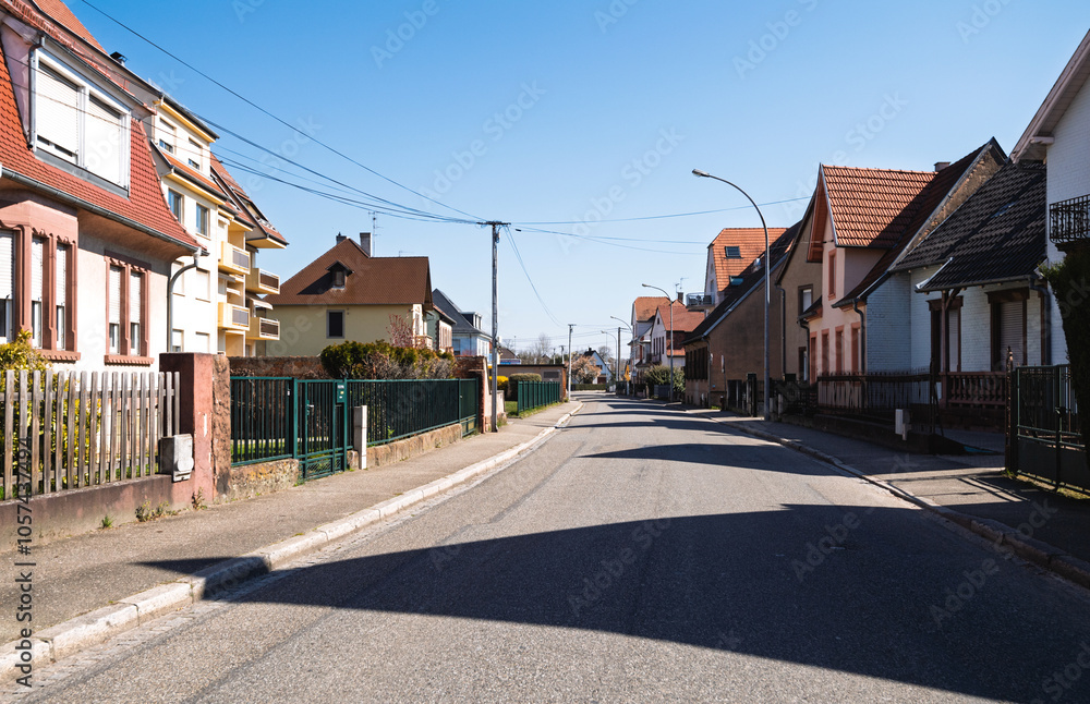 Empty residential street in a small French village with traditional houses, colorful facades, and fenced yards under a clear blue sky on a sunny day. Empty residential street in a small French village with traditional houses, colorful facades, and fenced yards under a clear blue sky on a sunny day.