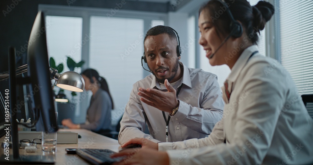 Obraz premium Call center office: Two multiethnic hotline operators in headsets discuss work, use computer having conversation with client on call. Technical support specialists working in online customer support.