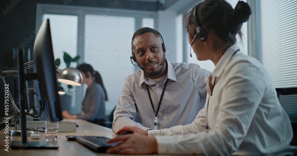 Fototapeta premium Call center office: Two multiethnic hotline operators in headsets discuss work, use computer having conversation with client on call. Technical support specialists working in online customer support.