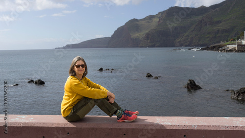 Woman poses for the camera as she sits on a concrete wall relaxing in the warm sun at the water's edge along the coast of the island of Madeira, Portugal; Canical, Madeira, Portugal photo