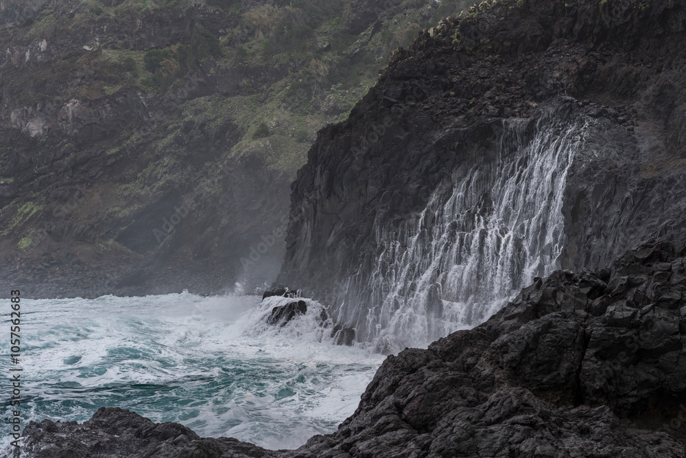 Water cascading from the rugged cliffs with a choppy ocean surf and mist along the coast of Madeira, Portugal; Seixal, Madeira, Portugal Water cascading from the rugged cliffs with a choppy ocean surf and mist along the coast of Madeira, Portugal; Seixal, Madeira, Portugal