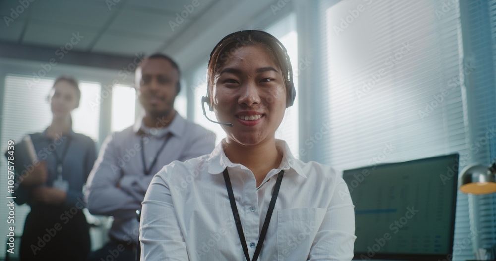Call center office: Close up portrait of friendly Asian female technical customer support specialist wearing headset smiling, looking at camera. Team of hotline operators standing in the background. Call center office: Close up portrait of friendly Asian female technical customer support specialist wearing headset smiling, looking at camera. Team of hotline operators standing in the background.