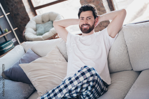 Photography Photo of attractive positive nice man wear pajama sitting on soft sofa hold arms
