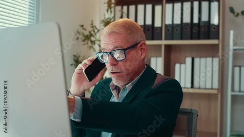 Businessman is sitting at his desk, talking on the phone and pointing at his computer screen. He is wearing a suit and glasses