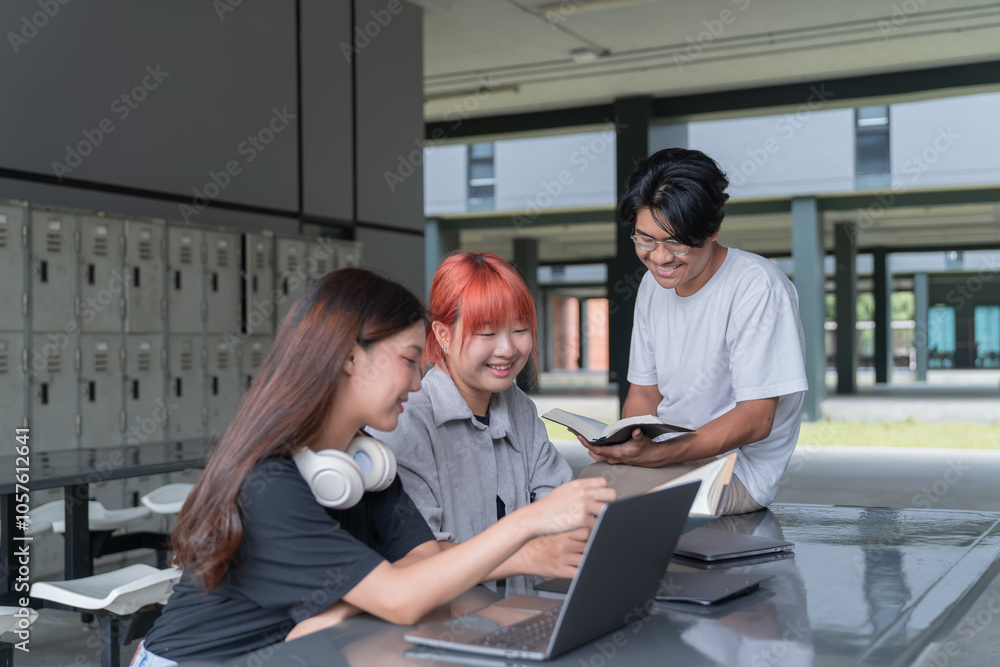 Three cheerful asian students are engaging in teamwork, utilizing a laptop and textbook to complete their assignment within the university campus Three cheerful asian students are engaging in teamwork, utilizing a laptop and textbook to complete their assignment within the university campus