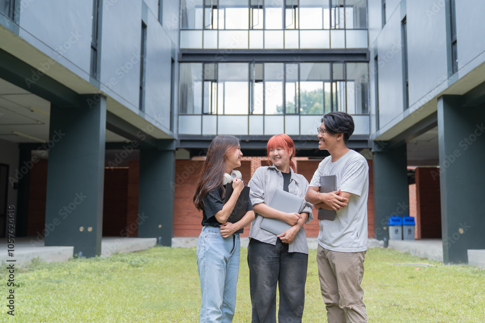 Three cheerful asian students are discussing and smiling on the lawn of their university campus, holding laptops and folders Three cheerful asian students are discussing and smiling on the lawn of their university campus, holding laptops and folders