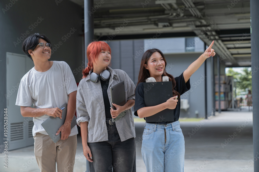 Obraz premium Three young students, holding laptops and clipboard, are standing together outdoors on a campus, with one student pointing towards something in the distance while engaging in a discussion
