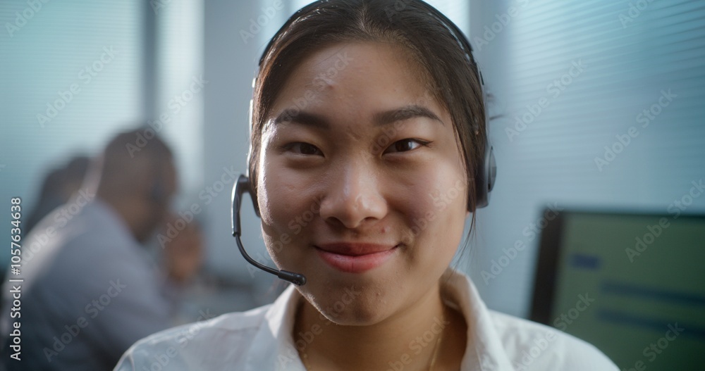 Close up of friendly Asian woman in headset working in call center office. Female technical customer support specialist or hotline operator sits at workplace, smiles and looks at camera. Portrait. Close up of friendly Asian woman in headset working in call center office. Female technical customer support specialist or hotline operator sits at workplace, smiles and looks at camera. Portrait.