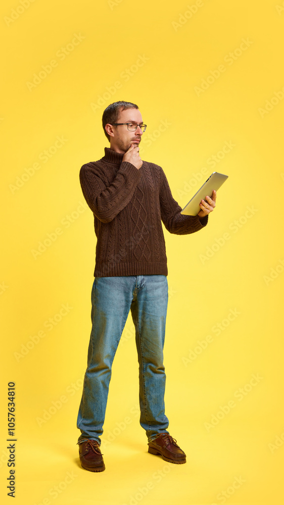 Thoughtful young man watching in tablet and choose presents or clothing online against yellow studio background. Concept of human emotions, beauty and fashion, business, shopping, technology. Thoughtful young man watching in tablet and choose presents or clothing online against yellow studio background. Concept of human emotions, beauty and fashion, business, shopping, technology.