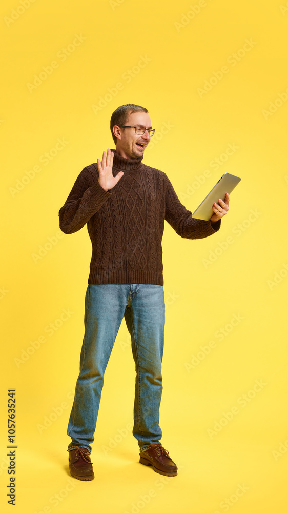 Young man dressed casual attire with tablet waving hello while holding meeting online against yellow studio background. Concept of human emotions, beauty and fashion, business, online. Young man dressed casual attire with tablet waving hello while holding meeting online against yellow studio background. Concept of human emotions, beauty and fashion, business, online.
