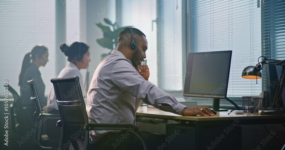 Multiethnic team of technical support operators working in modern call center office. African American helpdesk specialist talks on headset, uses computer, looks at camera providing customer service. Multiethnic team of technical support operators working in modern call center office. African American helpdesk specialist talks on headset, uses computer, looks at camera providing customer service.