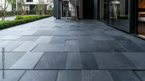 Modern outdoor patio with large dark slate tiles and reflective glass walls, surrounded by green landscaping in front of a contemporary building.