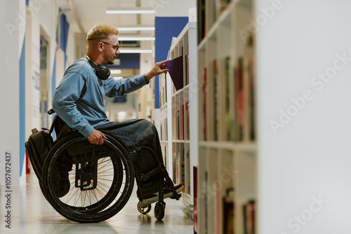 Young male student in wheelchair browsing library shelves for books while wearing headphones and casual attire. Focused individual engaging in academic environment with assistive device photo