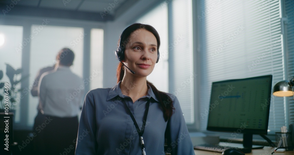 Call center office: Close up portrait of friendly Caucasian female technical customer support specialist wearing headset smiling, looking at camera. Team of hotline operators standing in background. Call center office: Close up portrait of friendly Caucasian female technical customer support specialist wearing headset smiling, looking at camera. Team of hotline operators standing in background.