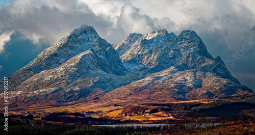 Cultivated land and settlement by snow covered mountains in a cloudy sunny day. Lofoten Islands, Northern Norway.