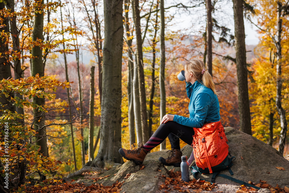 Female hiker relaxing on rock and drinking coffee in autumn woods with red backpack. Woman resting after successful hike Female hiker relaxing on rock and drinking coffee in autumn woods with red backpack. Woman resting after successful hike