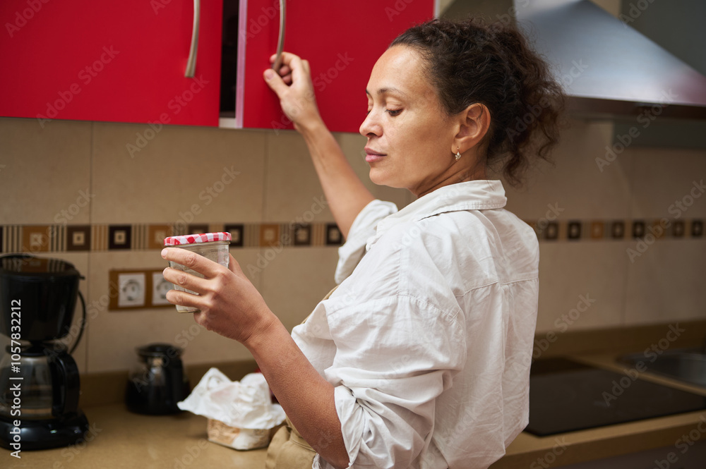 Woman in kitchen organizing pantry, reaching for cabinet while holding glass jar, showcasing domestic life, routine, and home organization in casual, modern interior setting.