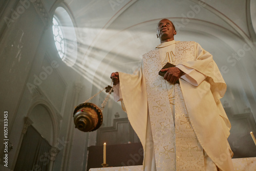 Low angle shot of mature African American priest in golden white robe swinging golden censer while besmoking with incense church