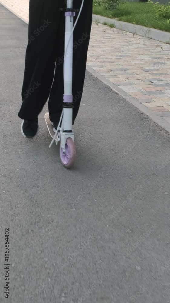 Vertical video. Close-up of schoolgirl rides her scooter on a tranquil path as she moves to school in a lively neighborhood in the morning Vertical video. Close-up of schoolgirl rides her scooter on a tranquil path as she moves to school in a lively neighborhood in the morning