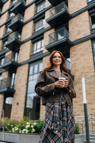 Vertical portrait of positive young woman savoring coffee break in urban environment, holding cup and sipping drink with smile, exuding joy and relaxation, standing on background of modern building. photo