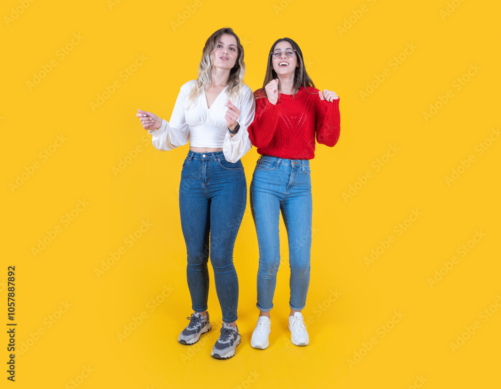 Fototapeta premium Energetic young two girls dancing, full body view caucasian 20s energetic young two girls dancing side by side waving hands in studio yellow background. Smiling happy two women listen music.