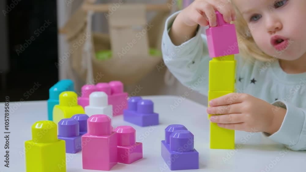 A little child toddler girl sits at a table and builds block towers out of a construction kit.