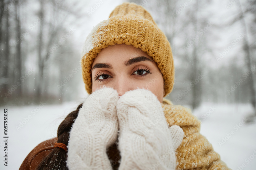 Close up portrait of young woman in winter outdoors looking at camera and blowing on hands with white fluffy mittens Close up portrait of young woman in winter outdoors looking at camera and blowing on hands with white fluffy mittens