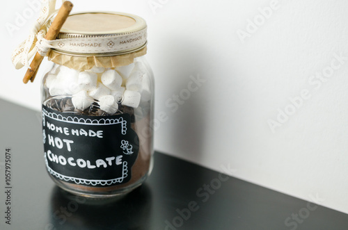 Homemade hot chocolate with marshmallows, in a jar with handmade label decorated with cinnamon stick, idea for a homemade gift, on a black table, white wall, expressive composition photo