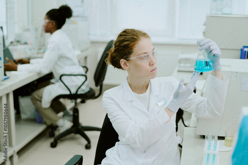 Scientist holding blue chemical solution in laboratory while a colleague working in background, both focused on tasks and using advanced equipment surrounded by scientific tools Scientist holding blue chemical solution in laboratory while a colleague working in background, both focused on tasks and using advanced equipment surrounded by scientific tools