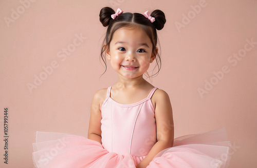 Adorable young Asian girl in pink ballet outfit smiling at camera. Studio portrait featuring double bun hairstyle with bows, tutu dress against pink backdrop