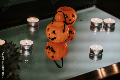 Miniature orange pumpkin buckets with jack-o'-lantern faces and tealight candles arranged on a reflective glass surface, creating a spooky Halloween ambiance.