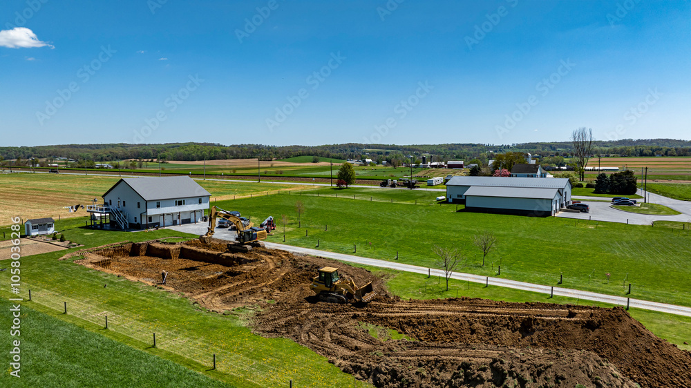 Heavy machinery works on a construction site near a house, surrounded by fields under a clear blue sky. The area is bustling with activity, indicating progress on new development.
