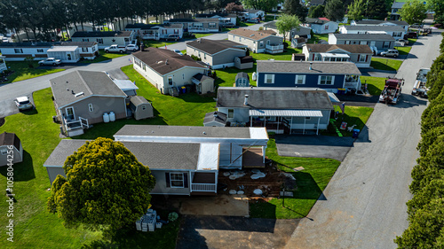 Fototapeta Naklejka Na Ścianę i Meble -  Aerial view shows mobile, manufactured homes lined along a tranquil street, surrounded by well-maintained lawns and trees. The sun casts a warm light, highlighting the neat layout and construction.