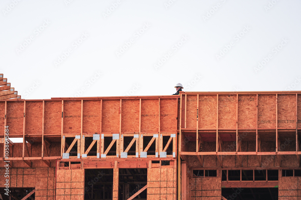 A construction worker wearing a safety helmet stands on the top level of a wooden-framed building under construction against a clear sky, showcasing modern architectural development A construction worker wearing a safety helmet stands on the top level of a wooden-framed building under construction against a clear sky, showcasing modern architectural development