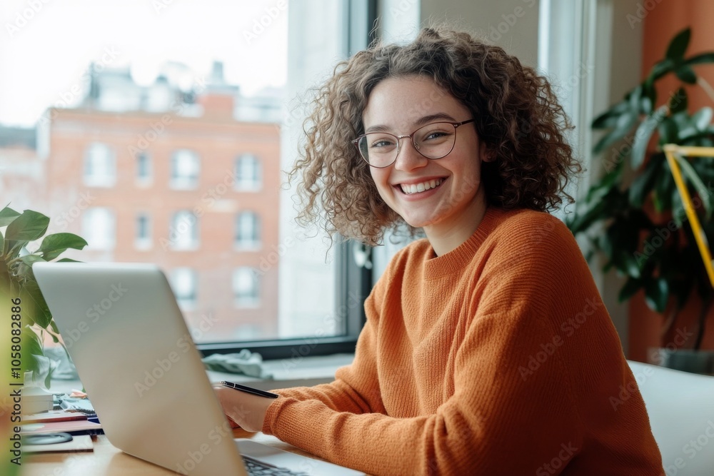 Smiling woman with curly light brown hair wearing glasses and orange sweater, sitting at desk with laptop, smartphone, and pen, looking into the camera. Bright room with natural light and green plants Smiling woman with curly light brown hair wearing glasses and orange sweater, sitting at desk with laptop, smartphone, and pen, looking into the camera. Bright room with natural light and green plants