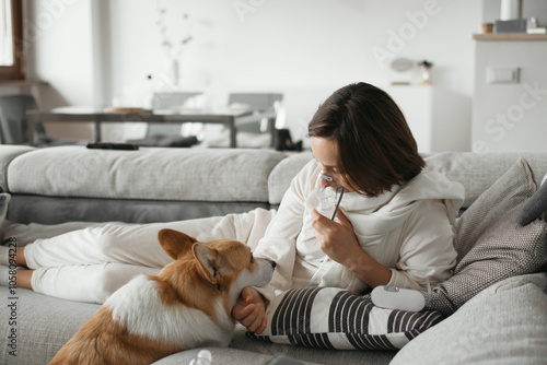 Young Woman Is Lying On The Sofa, Using A Nebulizer For Inhalation, While Petting Her Welsh Corgi Pembroke Dog