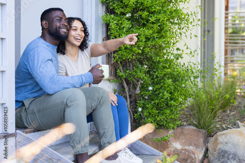 Smiling multiracial couple enjoying coffee on porch, pointing at something outside