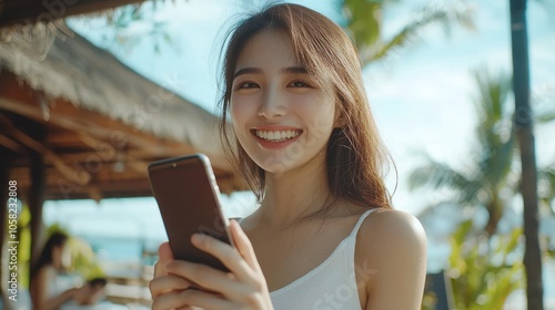 Smiling young Asian woman wearing white top holding a smartphone while looking to the camera, nature cafe background. photo