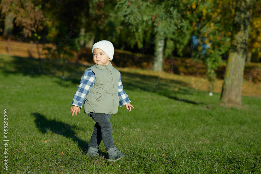 A young child stands on green grass in an autumn park, dressed warmly in a beanie, plaid shirt, and vest, looking curiously at the camera. The background is filled with fall foliage. A young child stands on green grass in an autumn park, dressed warmly in a beanie, plaid shirt, and vest, looking curiously at the camera. The background is filled with fall foliage.