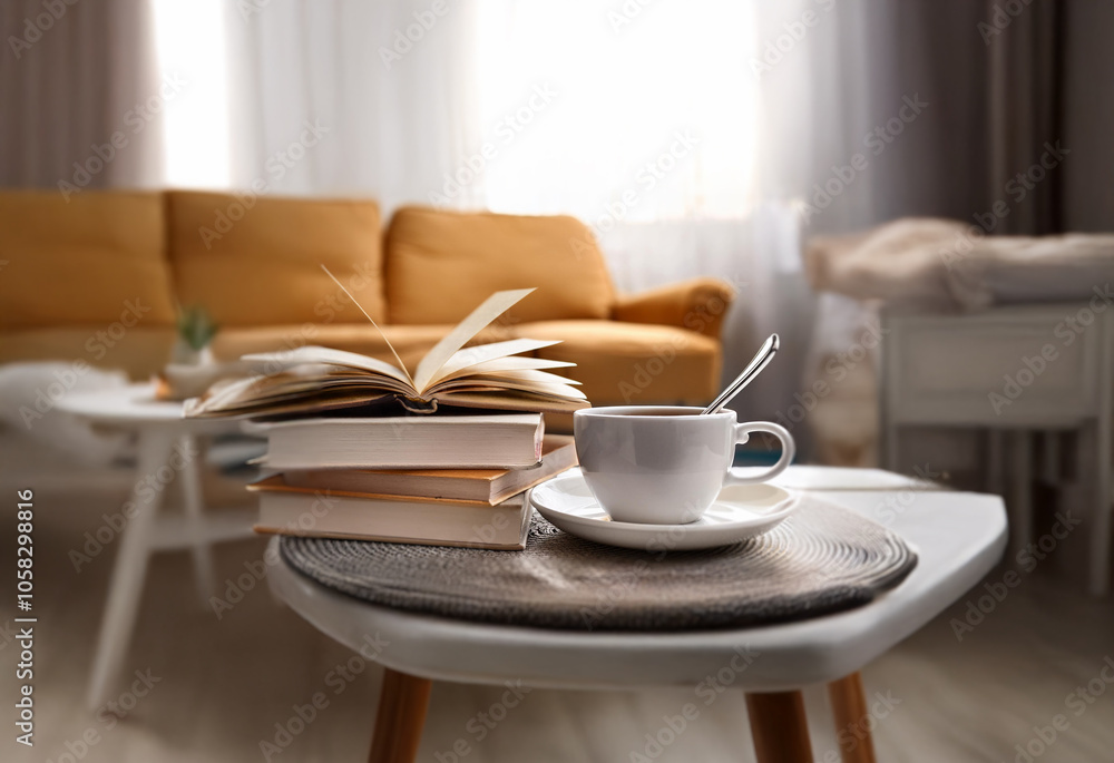 A cup of coffee and a stack of books rest on a small table in a living room with a yellow sofa. A cup of coffee and a stack of books rest on a small table in a living room with a yellow sofa.