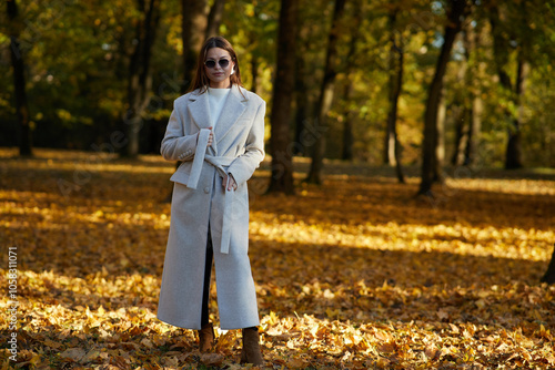 A stylish woman in a light beige coat stands confidently in an autumn park, surrounded by golden leaves. Sunlight filters through the trees, creating a warm, cozy seasonal atmosphere.