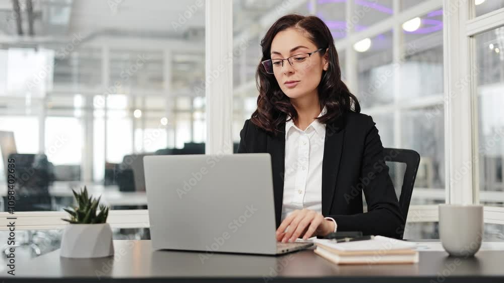 Focused businesswoman sits at her desk and puts on her glasses to work on her laptop. Woman is focused and productive. Female office worker starts work Focused businesswoman sits at her desk and puts on her glasses to work on her laptop. Woman is focused and productive. Female office worker starts work