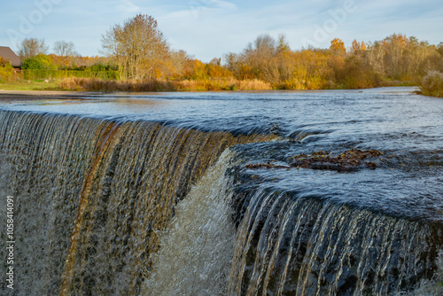 Beauty of Jagala Waterfall in Estonia, a popular natural attraction (Estonian - Jägala juga). The waterfall cascades over a rock ledge. In the background autumn trees with shades of orange and yellow. photo