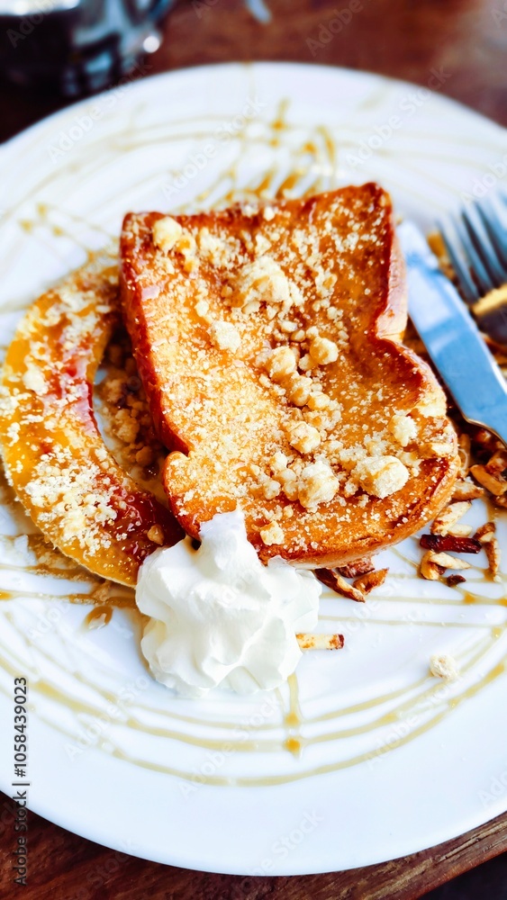 A plate of French toast topped with crumbs and a drizzle of caramel sauce, accompanied by a dollop of whipped cream and a side of nuts. A fork and knife rest beside it on the plate.
A plate of French toast topped with crumbs and a drizzle of caramel sauce, accompanied by a dollop of whipped cream and a side of nuts. A fork and knife rest beside it on the plate.