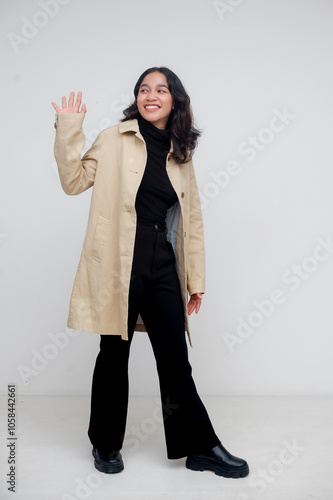 Whole body photo of a fashionabel young asian woman wearing a classic beige trench coat over a black turtleneck top, waving hi to a friend. White background. photo