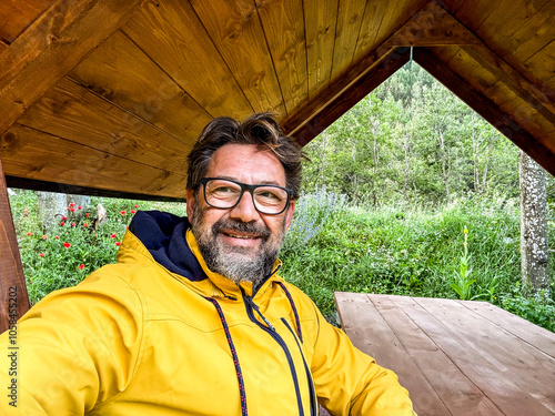 Friendly middle-aged man in trendy yellow jacket sits in a park, happily engaging with his smartphone camera for a social media video call selfie, capturing natural joy