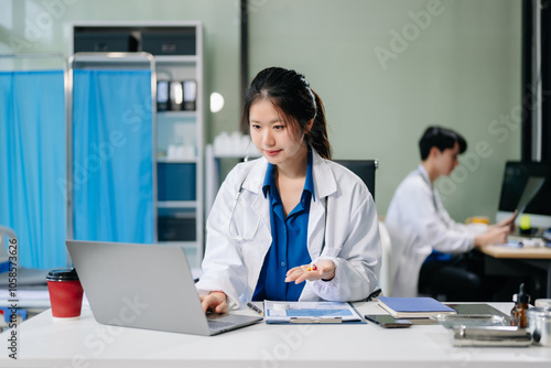 Confident young asian female doctor in white medical uniform sit at desk working on computer.