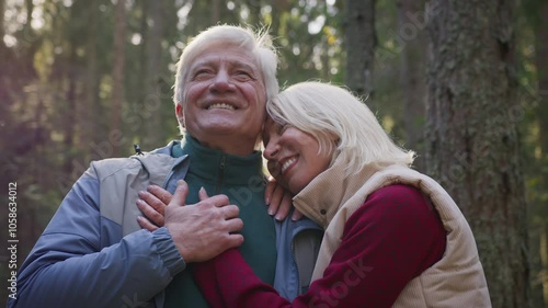 Senior elderly couple stay in sunny forest, hugging and enjoying leisure together. Tender love of old married couple embraces during stroll through the forest, smiling and admiring nature. Retirement