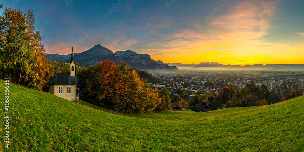 Obraz premium Panorama Sunset in the Rhine Valley, with a tree and a chapel,blue sky over the city of Dornbirn, meadow and fields. with Swiss mountains in the background. afterglow, interesting colored veil clouds