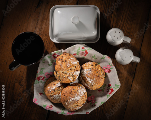 Classic farm country bread buns in a cute basket next to a mug, butter, salt, pepper. These homemade rustic rolls balls are made of organic wholemeal multigrain wheat flour from sourdough recipe.